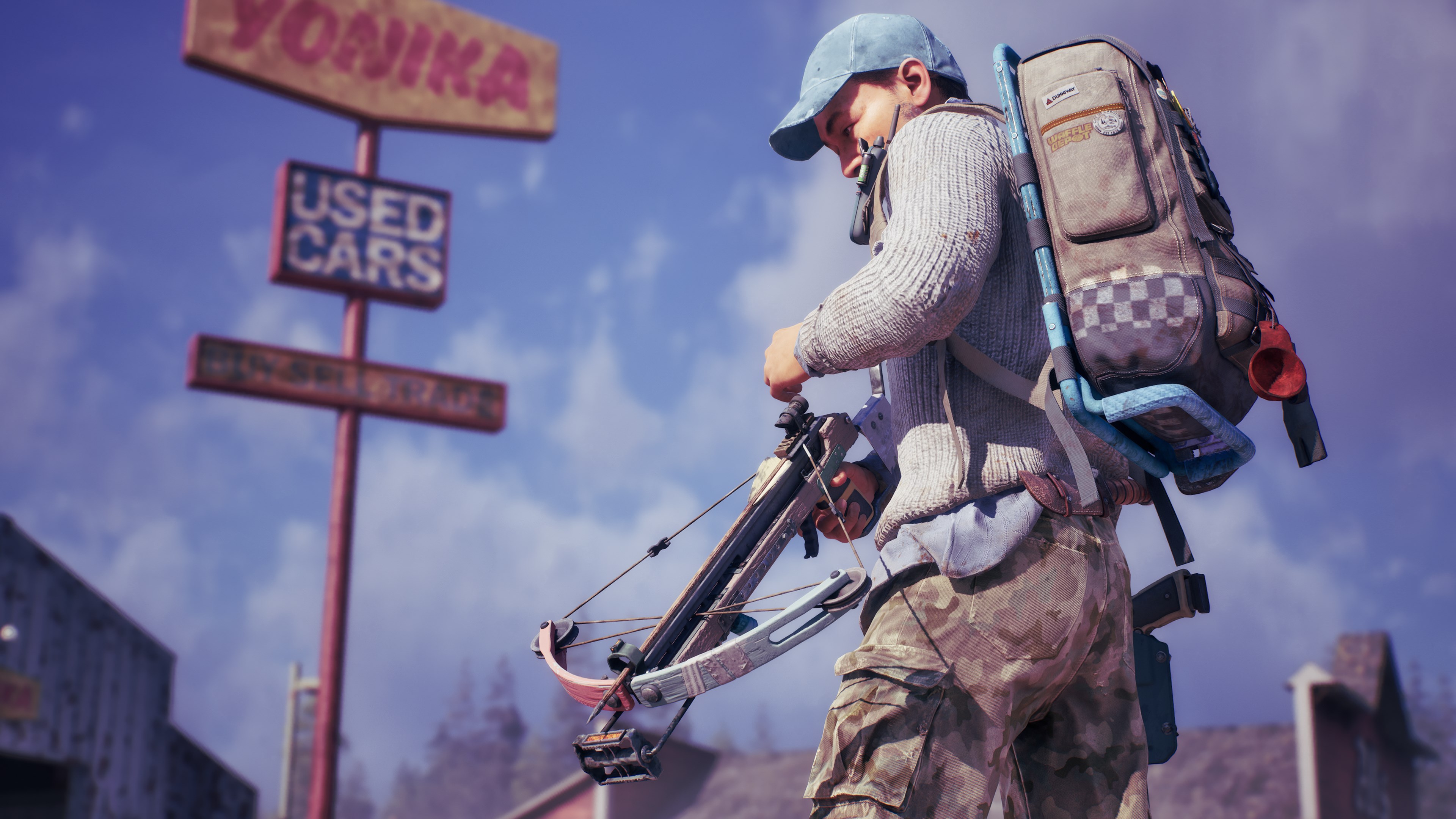 A survivor in a cap and knitted sweater handles a crossbow beside a derelict used car lot sign, a loaded backpack strapped to their shoulders