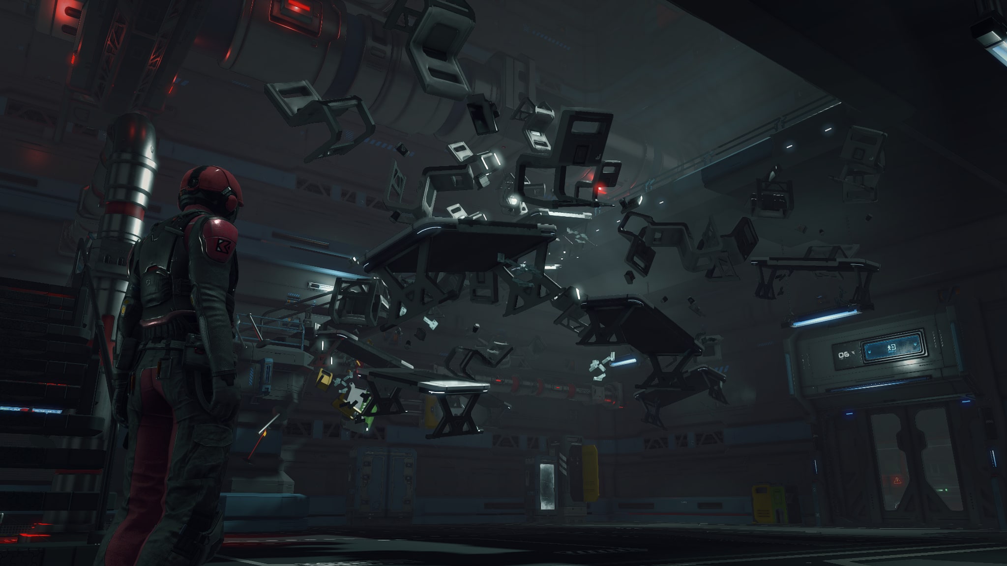 A player character in a red suit stands in a dark industrial hall while tables, chairs and debris float in mid-air around a door marked 06