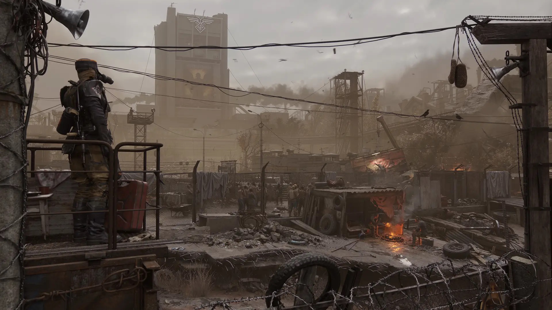 An armed guard on a walkway overlooks a wired, dust-hazed surface camp of scrap shelters and loudspeakers, with the Novoreich emblem mounted on a concrete tower behind it.