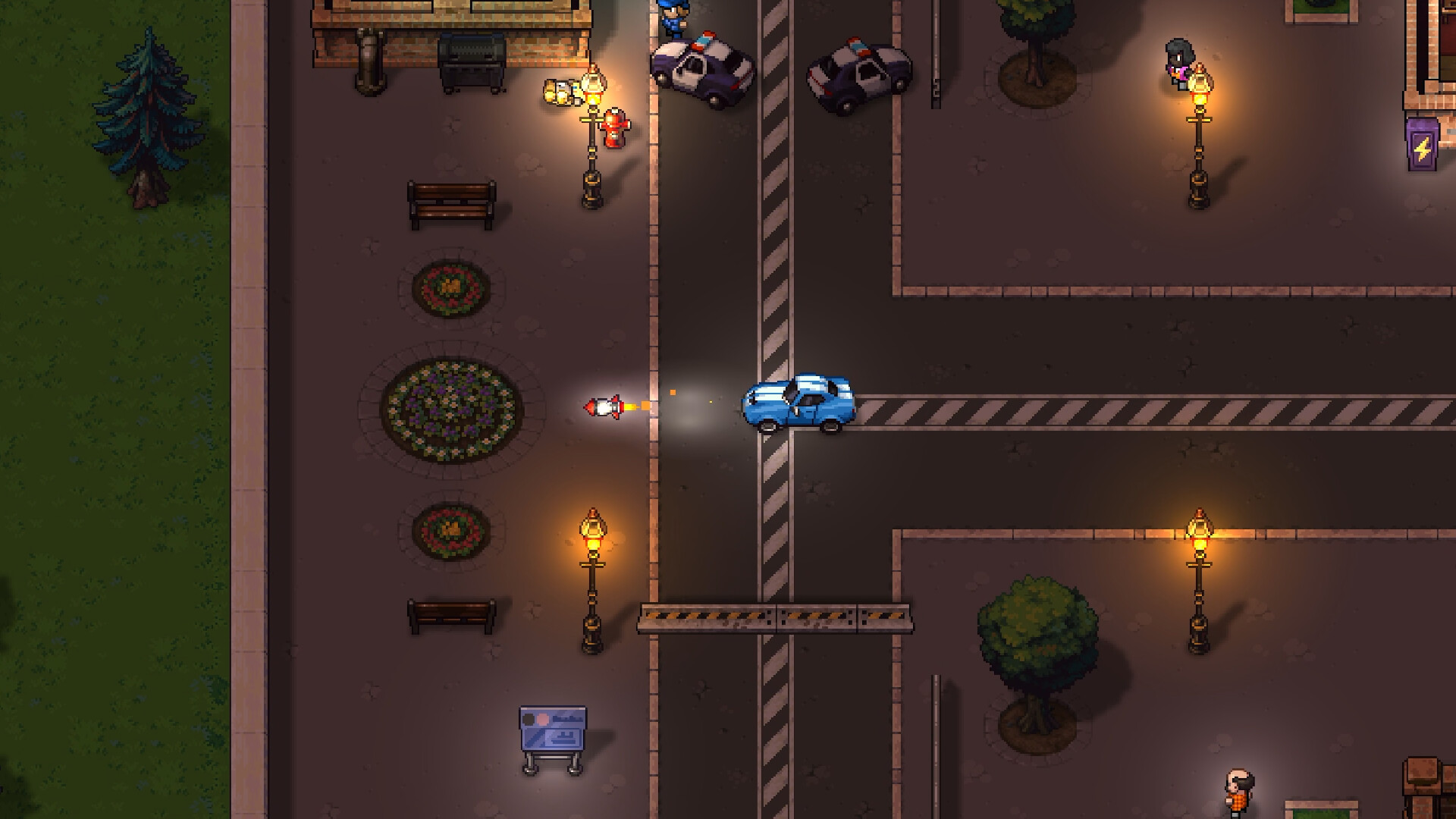 A rocket streaks across a night intersection in Streets of Rogue 2 toward a moving blue car, while two police cruisers sit parked at the kerb beside flower beds, benches and a construction barrier