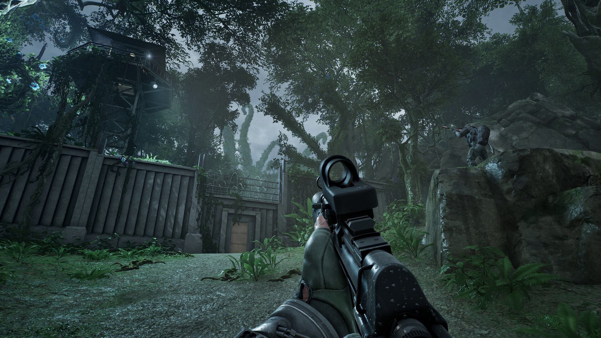 First-person view of a rifle fitted with an optic while approaching a walled compound topped with barbed wire and a raised watchtower in the jungle, an opposing Contractor aiming from a rock outcrop on the right