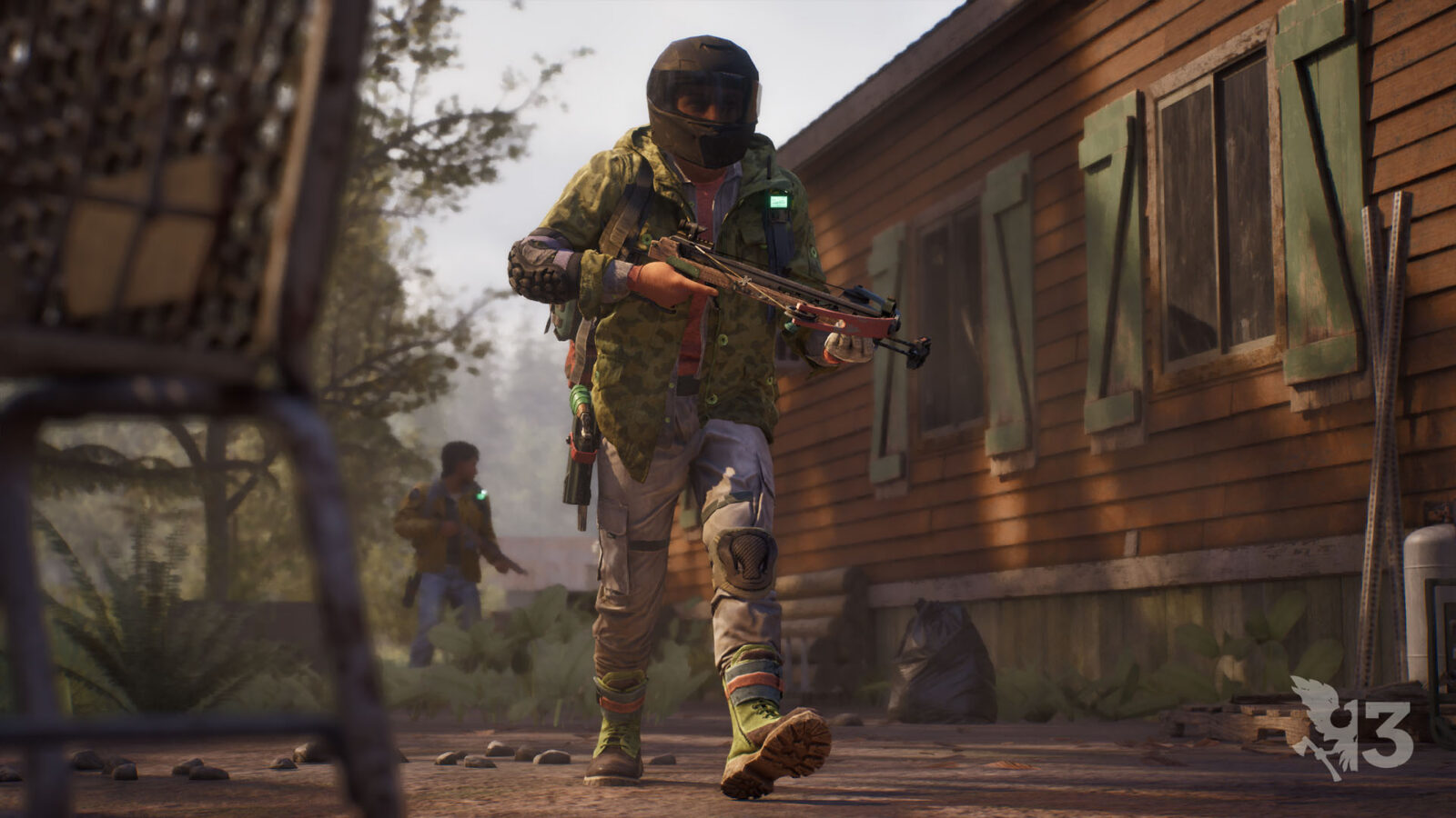 A survivor in a full-face helmet and a patched camouflage jacket carries a compact crossbow past a wooden house, with a second survivor approaching through the trees behind.